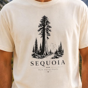 May include: A cream-colored short-sleeve t-shirt featuring a black graphic print of a large sequoia tree in a forest setting with mountains in the background. The text "SEQUOIA" is printed below the tree, followed by "Walk Among Giants".