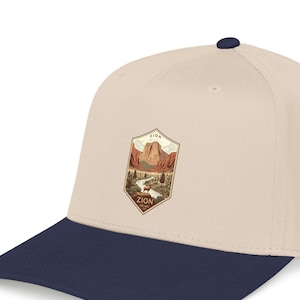 May include: A beige baseball cap with a navy blue peak and a navy blue button. The cap has a patch with a landscape illustration of Zion National Park, including the text "ZION".