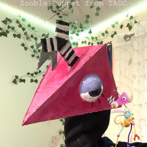 May include: A handmade Zooble Puppet from TADC, featuring a pink triangular head with a large, round eye and a black and white striped detail. The puppet has a whimsical, cartoonish design.