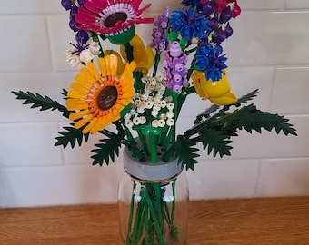 May include: A colourful bouquet of Lego flowers in a clear glass jar. The flowers include pink, purple, yellow, and blue blooms with green stems and leaves.