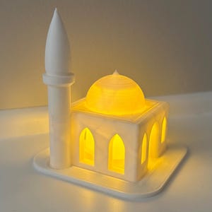 May include: A white, illuminated mosque model with a minaret and dome. The miniature building has arched windows and a warm, yellow glow emanating from within, creating a soft, inviting ambiance. The model sits on a white base.