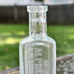 Könnte beinhalten: Klare Glasflasche mit geprägtem Text: "DR HARVEY'S ALPINE BALSAM A.W. DOBBS ITHACA NY". Die Flasche hat eine rechteckige Form mit schmalem Hals und flachem Boden. Der Hintergrund ist unscharf.