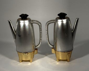 May include: Two silver-colored metal coffee pots with ribbed handles and black knobs on the lids. Each pot features a gold-colored base with three short legs and a spout extending from the side of the main cylindrical body.