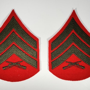Może przedstawiać: Dwie czerwono-zielone naszywki z oznaczeniem stopnia sierżanta. Każda naszywka ma trzy ciemnozielone szewrony i skrzyżowane karabiny na czerwonym tle. Naszywki są prawdopodobnie do munduru wojskowego.