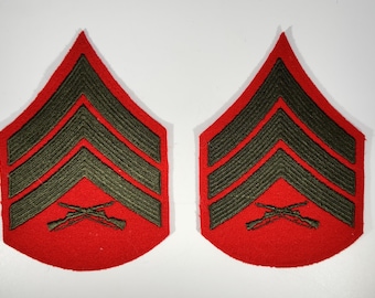 May include: Two red and green Sergeant rank insignia patches. Each patch features three dark green chevrons and crossed rifles on a red background. The patches are likely for a military uniform.