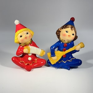 Puede incluir: Dos figuras de payasos de cerámica. Uno viste de rojo y toca un acordeón, el otro de azul toca un instrumento amarillo. Ambos tienen caras y sombreros pintados. Las figuras están sentadas.