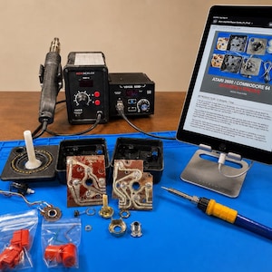 May include: A workspace with electronic repair tools and components. Includes a soldering iron, heat gun, and a tablet displaying a repair guide for Atari 2600 and Commodore 64 consoles. Various small parts and a game controller are also present.