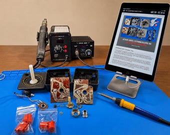 May include: A workspace with electronic repair tools and components. Includes a soldering iron, heat gun, and a tablet displaying a repair guide for Atari 2600 and Commodore 64 consoles. Various small parts and a game controller are also present.