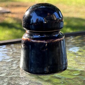 May include: A dark blue ceramic insulator with a glossy finish. The antique piece has a rounded top and a flared base. The object is sitting on a glass surface, with a blurred green background.