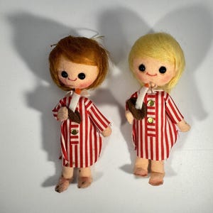Puede incluir: Dos muñecas vintage de fieltro con caras redondas, ojos negros y mejillas rosadas. Tienen cabello rojo y rubio y visten vestidos a rayas rojas y blancas con botones dorados. Cada muñeca sostiene un objeto marrón y una vela blanca.