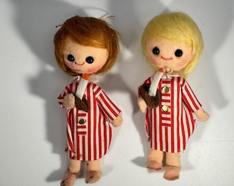 May include: Two vintage felt dolls with round faces, black eyes, and rosy cheeks. They have red and blonde hair, and wear red and white striped dresses with gold buttons. Each doll holds a brown object and a white candle.
