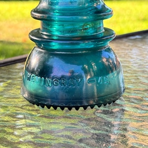 Può includere: Un isolatore elettrico in vetro color verde acqua con una parte superiore arrotondata e una base larga. Il vetro presenta la scritta in rilievo HEMINGRAY - 42. Il bordo inferiore dell'isolatore presenta una serie di piccole tacche equidistanti.