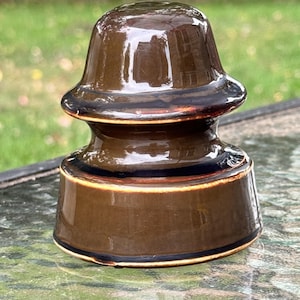 May include: A brown ceramic insulator with a glossy finish. The antique piece has a rounded top and a cylindrical base, with dark brown and black accents. The insulator is likely from an electrical or telephone system.