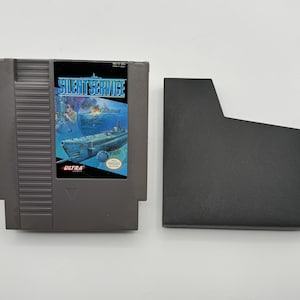 May include: A gray Nintendo Entertainment System (NES) game cartridge with the title "Silent Service" printed on the label. The game features a submarine and a blue ocean background. The cartridge is next to a gray plastic case.