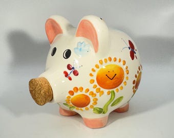 May include: A ceramic piggy bank in a cream color with a cork stopper in the snout. The body is decorated with hand-painted orange sunflowers featuring smiling faces, green stems, and small red and blue butterflies. The ears and feet are pink.