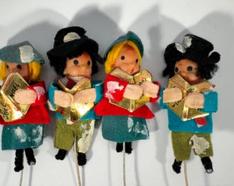 May include: Four vintage Christmas carolers on sticks. Each figure wears a hat, coat, and pants in various colors. They hold a golden book with the word "NOEL" on it. The figures have painted faces and black shoes.