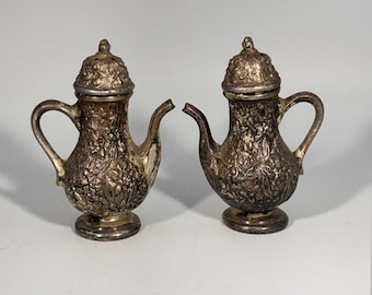 May include: A pair of small, metallic silver-toned shakers shaped like miniature teapots. Each piece features an intricate, embossed floral pattern covering the body and lid, a curved handle, a short spout, and a circular pedestal base.