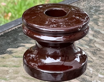 May include: A glossy, brown ceramic insulator with a rounded top and base, connected by a cylindrical midsection. The top features a central hole. The object is likely vintage and sits on a glass surface.