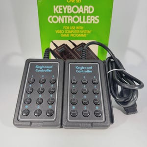 Può includere: Un set di due controller a tastiera rettangolari grigio scuro con pulsanti circolari neri numerati da zero a nove, più simboli asterisco e cancelletto. I controller presentano la scritta Keyboard Controller e una scatola verde.