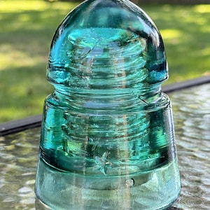 Pode incluir: Um isolador de vidro turquesa vintage com um topo arredondado e um design em forma de estrela. O isolador tem um interior roscado e um acabamento liso e brilhante. Está sobre uma superfície reflexiva.