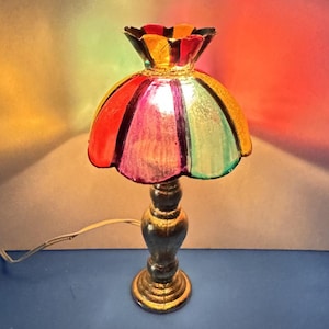 May include: A table lamp featuring a scalloped, multi-colored glass shade with panels of red, purple, green, and yellow. The lamp has a dark, textured, metallic-finish base with a curved, pedestal-style stem and a white electrical power cord.