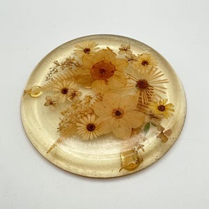 Puede incluir: Un posavasos redondo de resina transparente con un tinte dorado, que presenta una disposición de flores secas amarillas y naranjas. Las flores incluyen margaritas y flores más grandes, creando un diseño botánico natural. El posavasos tiene una superficie lisa y un ligero brillo.