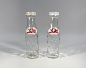 May include: Two vintage Pepsi-Cola glass bottles. The clear bottles have a diamond pattern and white caps. Each bottle has a red, white, and blue Pepsi-Cola logo. The bottles are empty and appear to be in good condition.