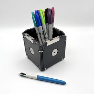May include: A black, cube-shaped pencil holder made from recycled floppy disks. The holder is filled with colorful markers. A blue pen is lying on a white surface in front of the holder.