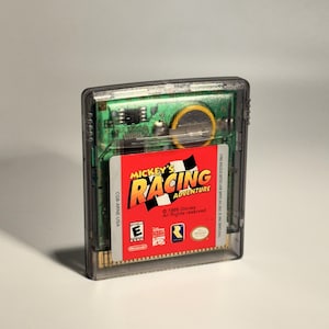 May include: A clear plastic Game Boy Color cartridge featuring "Mickey's Racing Adventure." The label is red with the game's title in yellow and white, and includes the ESRB rating. The cartridge's internal circuit board is visible.