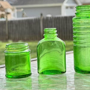 May include: Three green glass bottles of varying shapes and sizes. The bottles are empty and have a vintage appearance. The bottles are sitting on a reflective surface, with a blurred background of a fence and greenery.