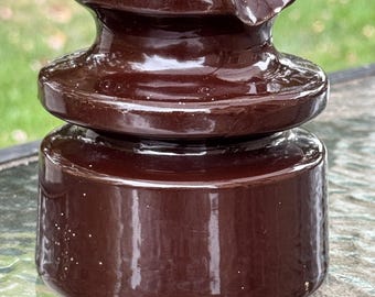 May include: A brown ceramic insulator with a glossy finish. The insulator has a rounded top with two white sections and a cylindrical base. It is designed to insulate electrical wires and is likely vintage.