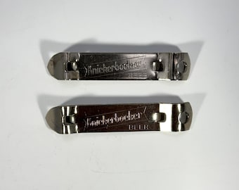 May include: Two silver-toned metal bottle openers featuring a flat, rectangular design with a pointed end and a circular hole at the opposite side. Each opener is embossed with the text Knickerbocker BEER in a stylized font across the center.
