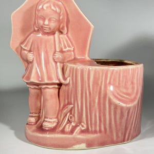 May include: A vintage pink ceramic planter featuring a young child holding an umbrella, standing next to a tree trunk. The planter has a textured finish and a brown interior. A charming decorative piece.
