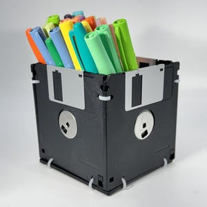 May include: A black desk organizer made from repurposed floppy disks, filled with a variety of colorful pens. The organizer is square-shaped, with the metal disk covers visible on the sides. The pens are in shades of orange, yellow, green, blue, and purple.