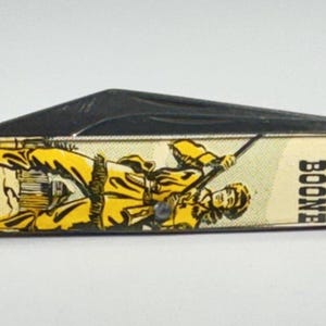 May include: A folding pocket knife with a cream-colored handle featuring a yellow and black illustration of a figure holding a rifle. The text DANIEL BOONE is printed in black on the right side of the handle. The knife has a metallic blade.