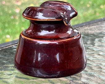 May include: A glossy, dark brown ceramic insulator with a rounded base and a top section with two curved protrusions. The antique piece has a smooth, reflective surface, suggesting its age and use in electrical systems.