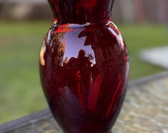 May include: A deep ruby red glass vase with a smooth, glossy finish. The vessel features a rounded, bulbous body that tapers toward a flared rim and a circular base, reflecting light across its translucent, dark crimson surface.