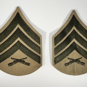 May include: Two tan and olive green military rank insignia patches. Each patch features three chevrons and crossed rifles, indicating a specific rank within the armed forces. The patches are likely made of fabric and are designed to be sewn onto a uniform.