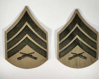 May include: Two tan and olive green military rank insignia patches. Each patch features three chevrons and crossed rifles, indicating a specific rank within the armed forces. The patches are likely made of fabric and are designed to be sewn onto a uniform.