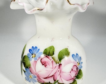 May include: A white milk glass vase with a ruffled, gold-trimmed rim. The body features a hand-painted floral design with two large pink roses, green leaves, and small blue flowers. The neck has a subtle, repeating cross-stitch pattern.