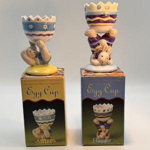 May include: Two ceramic egg cups from the Muffy VanderBear Collection. One features a bunny holding a blue and white egg cup, the other a bunny holding a purple and yellow egg cup. Each sits atop a box labeled "Egg Cup".
