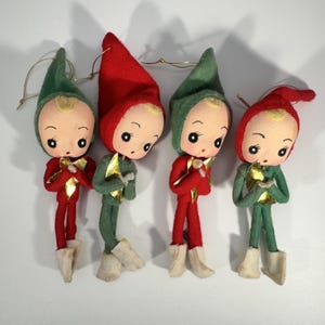 May include: Four vintage Christmas elf ornaments. Each elf has a fabric head with painted features, wearing a red or green outfit with a matching hat. Gold accents adorn the outfits, and they have cream-colored boots. Each ornament has a gold string for hanging.