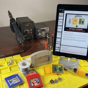 May include: A yellow work mat with various electronic repair tools, including a soldering iron, a heat gun, and a tablet displaying a battery replacement guide. Several video game cartridges and components are also present.