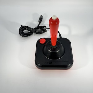 Puede incluir: Mando de juego negro con un mango rojo y un botón rojo. El mando tiene una base negra y una punta blanca en el mango. Un cable negro con un enchufe está conectado al mando.