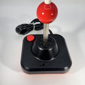 Puede incluir: Un joystick de videojuego negro con un mango esférico rojo rematado con un pequeño botón blanco. La base cuenta con un botón circular rojo a la izquierda y un cable negro en la parte posterior. El texto BASE y STICK está impreso al frente.