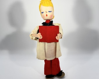 May include: A vintage doll with a yellow yarn hairstyle, blue eyelashes, and rosy cheeks, holding a red felt square. The doll wears a cream-colored coat, red pants, and black shoes. The doll's outfit includes a black bow tie.