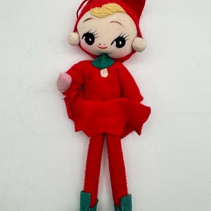 May include: A vintage doll with a red outfit, including a dress, hat, and tights. The doll has a painted face with large eyes, rosy cheeks, and blonde hair. It wears green shoes and has a pink hand. A charming collectible.