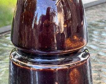 May include: A dark brown, glossy ceramic electrical insulator with a two-tiered, rounded dome shape. The surface is smooth and reflective, showing light reflections and a cylindrical base section topped by a smaller, rounded cap.