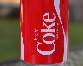 May include: A clear glass tumbler featuring a red wrap-around label with the white text Enjoy Coke and a registered trademark symbol. The glass has a cylindrical upper section and a slightly flared base, showcasing a classic beverage branding.