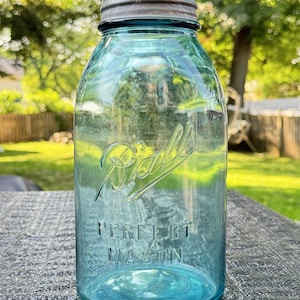 May include: A vintage aqua blue glass mason jar with a metallic screw-top lid. The front of the jar features the embossed cursive text Ball and the block letter text PERFECT MASON. The glass has a translucent blue tint and a cylindrical shape.